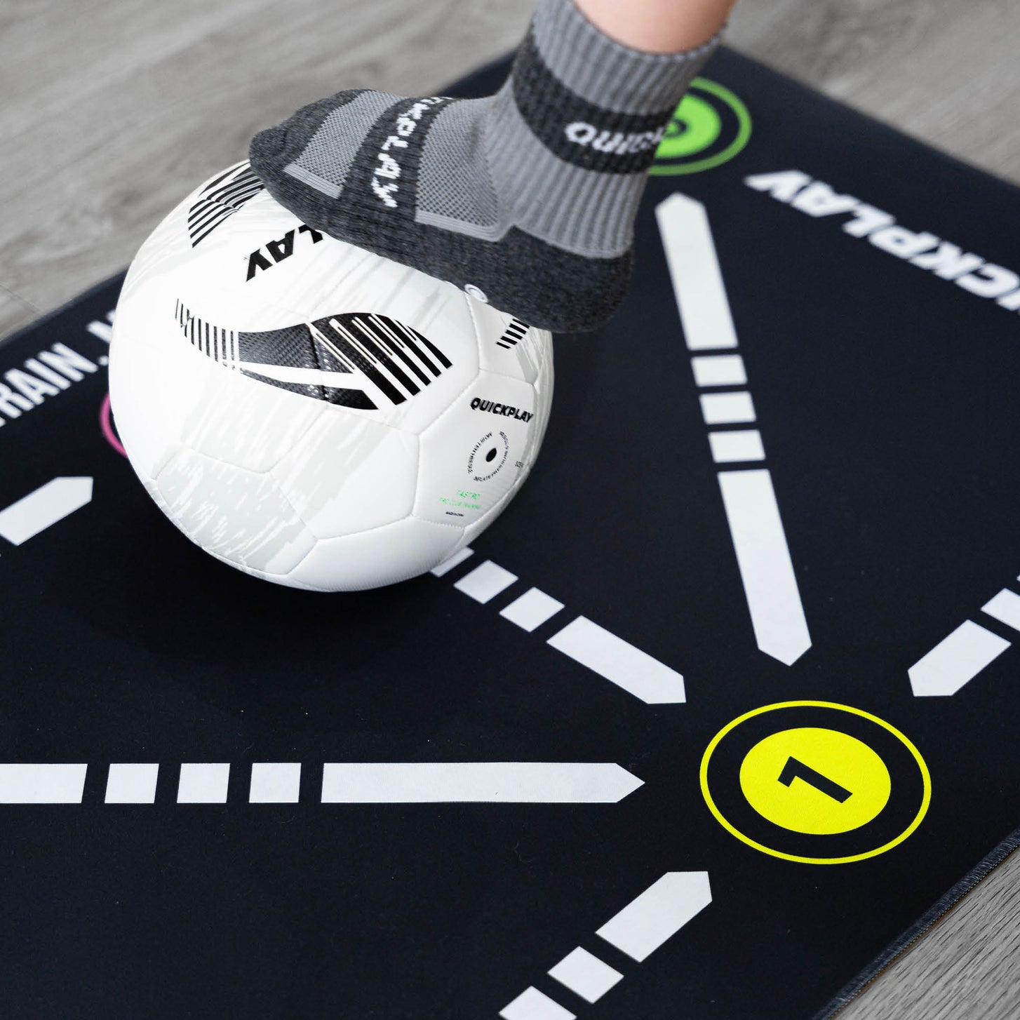 Primo piano del tappetino da calcio per il controllo palla, piede con una calza grip QUICKPLAY su una palla bianca accanto al numero 1 giallo.