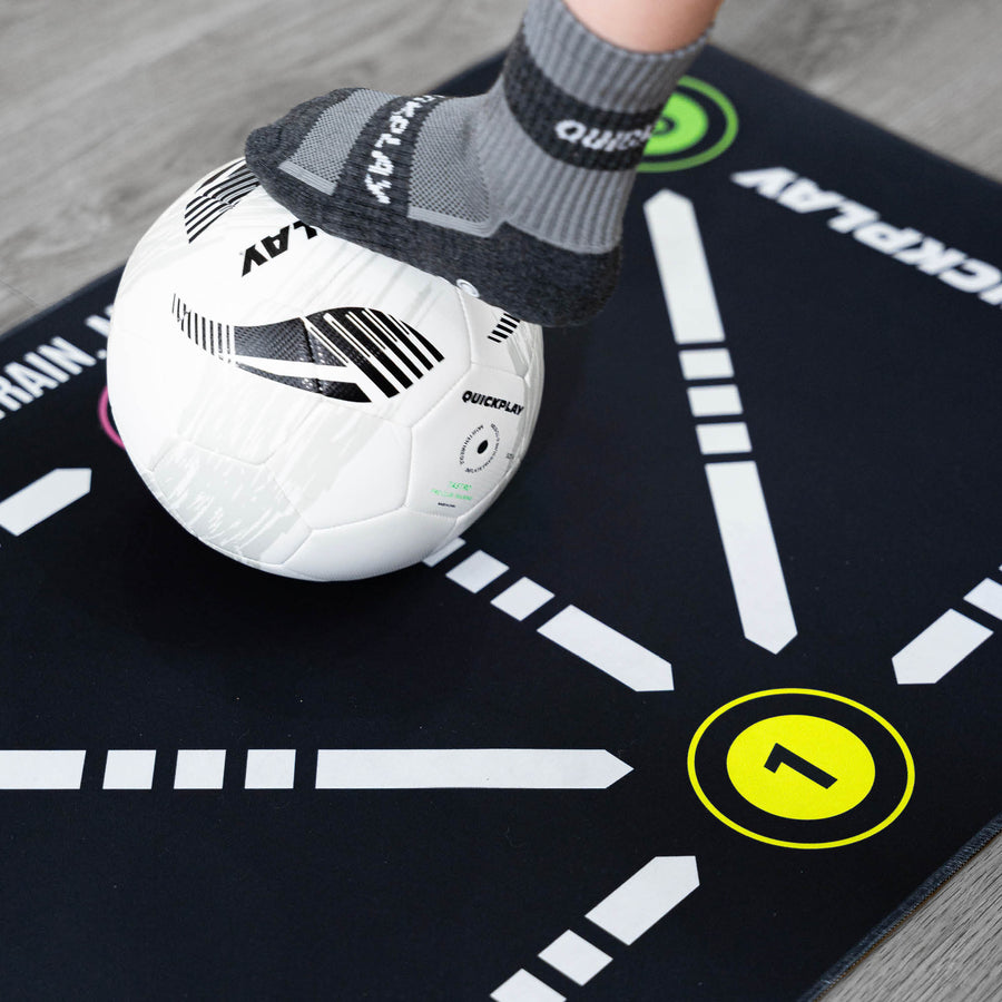Primo piano del tappetino da calcio per il controllo palla, piede con una calza grip QUICKPLAY su una palla bianca accanto al numero 1 giallo.