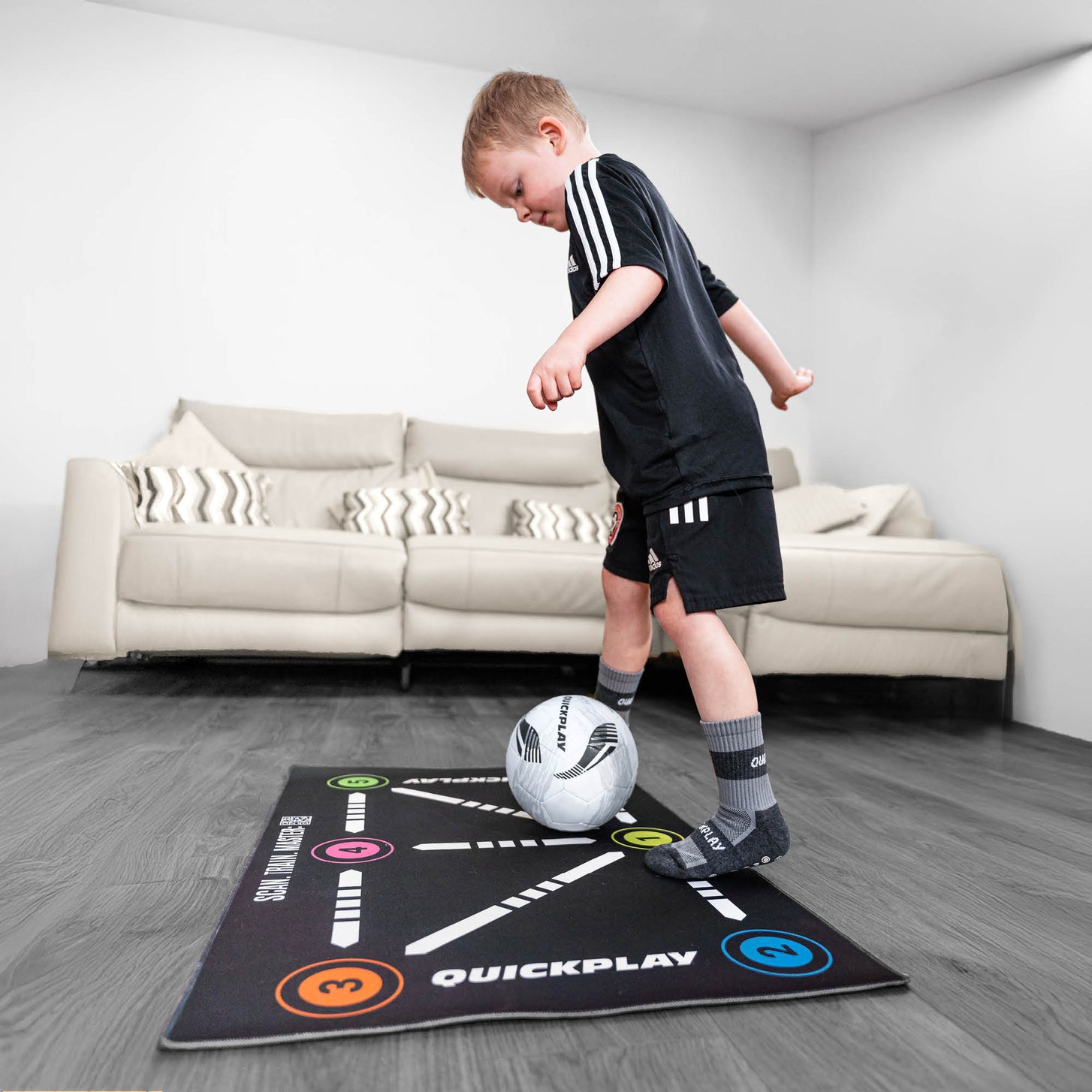 Bambino che si esercita a controllare la palla sul tappetino da calcio per il controllo palla su un pavimento in legno, con il divano dietro.