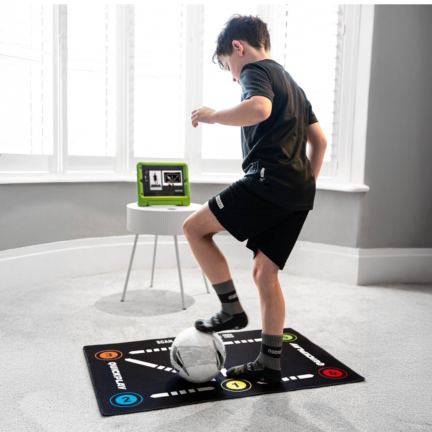 Bambino che fa un esercizio tecnico sul tappetino da calcio per il controllo palla su un tappeto, con un tablet che mostra un video vicino alla finestra.