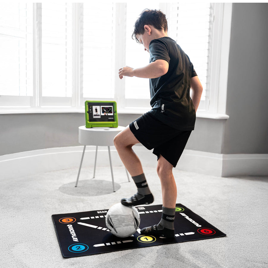 Bambino che fa un esercizio tecnico sul tappetino da calcio per il controllo palla su un tappeto, con un tablet che mostra un video vicino alla finestra.