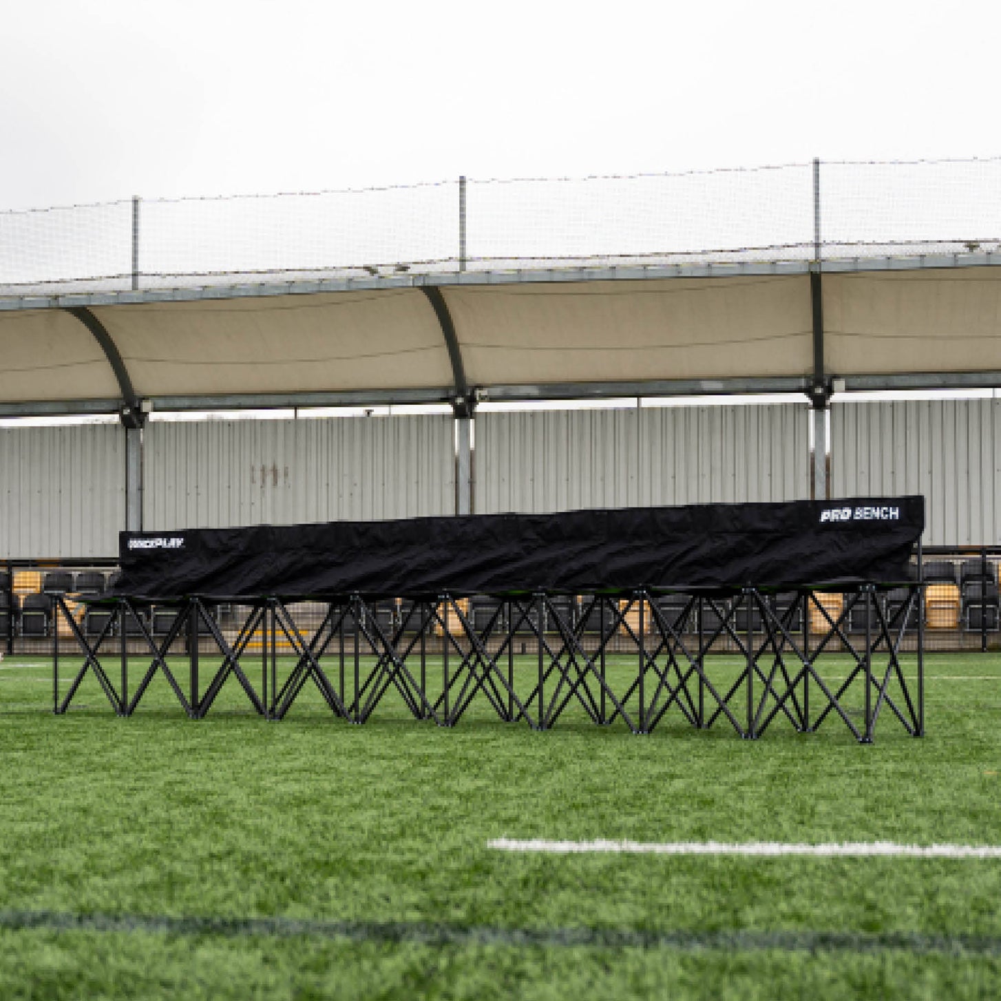 Lunga panchina PRO Bench 9 posti nera aperta su erba sintetica davanti a una tribuna coperta.