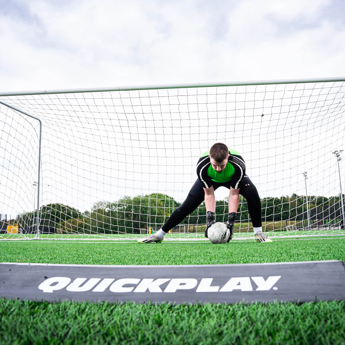 Portiere che raccoglie una palla bassa davanti alla porta, visto oltre il deviatore a rampa per l'allenamento dei portieri in primo piano.