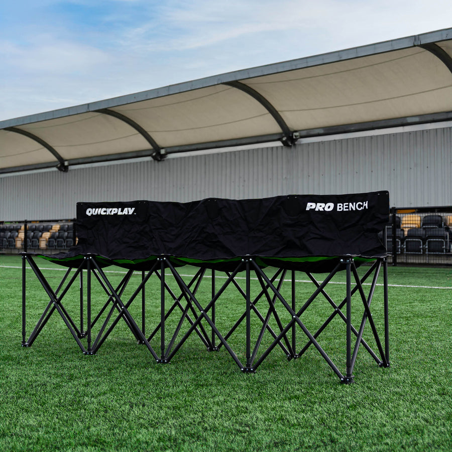 Panchina PRO Bench 4 posti nera aperta su erba sintetica davanti a una tribuna coperta di uno stadio.