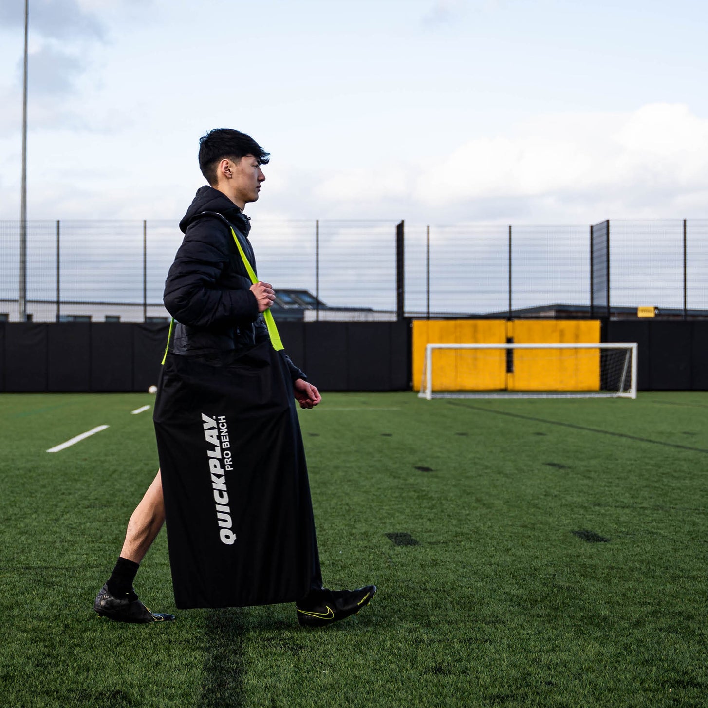 Giocatore che cammina su un campo con la panchina PRO Bench 4 posti riposta in una borsa nera QUICKPLAY PRO BENCH in spalla.