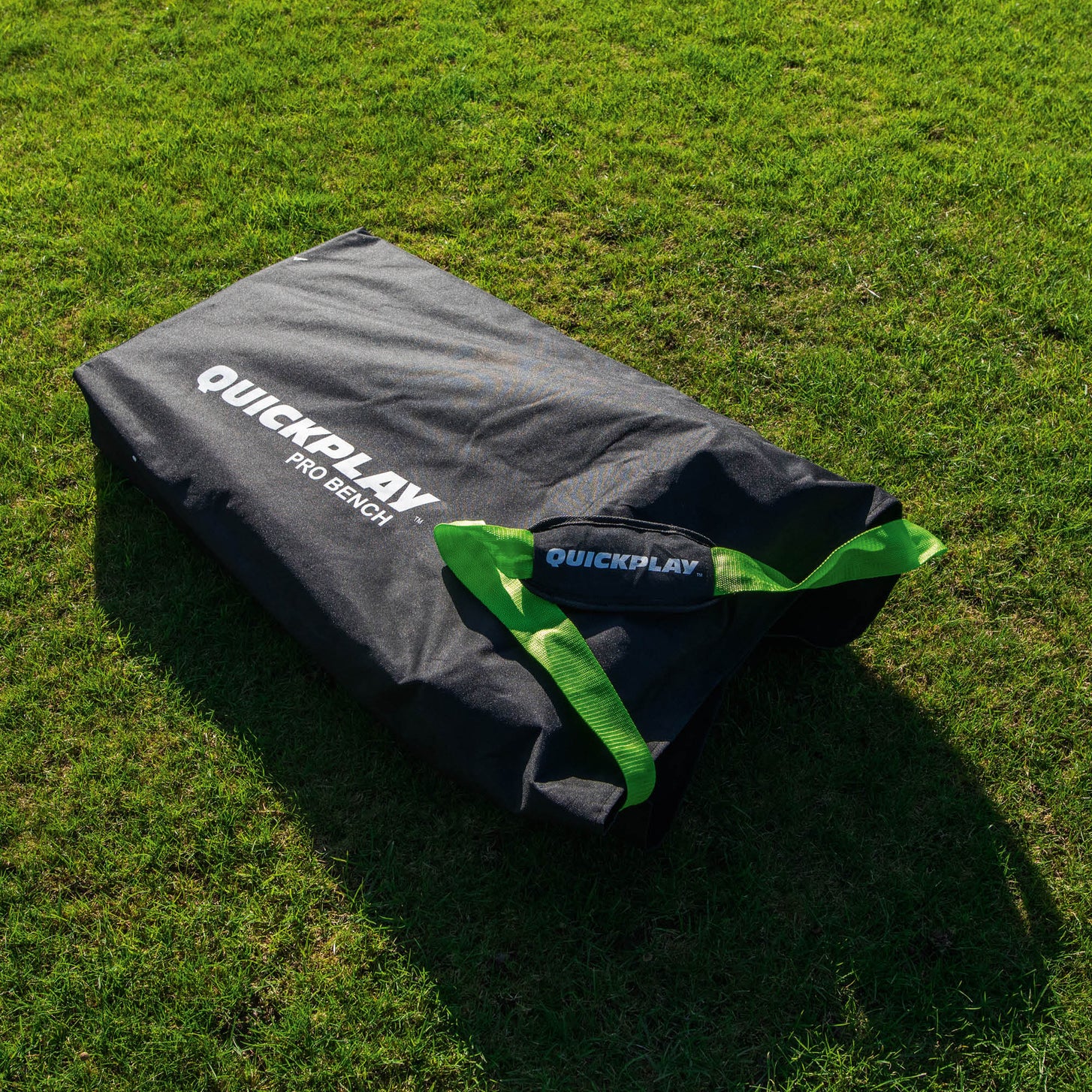 Borsa per il trasporto della panchina PRO Bench 4 posti, nera con la stampa QUICKPLAY PRO BENCH e cinghie verdi, distesa sull'erba al sole.