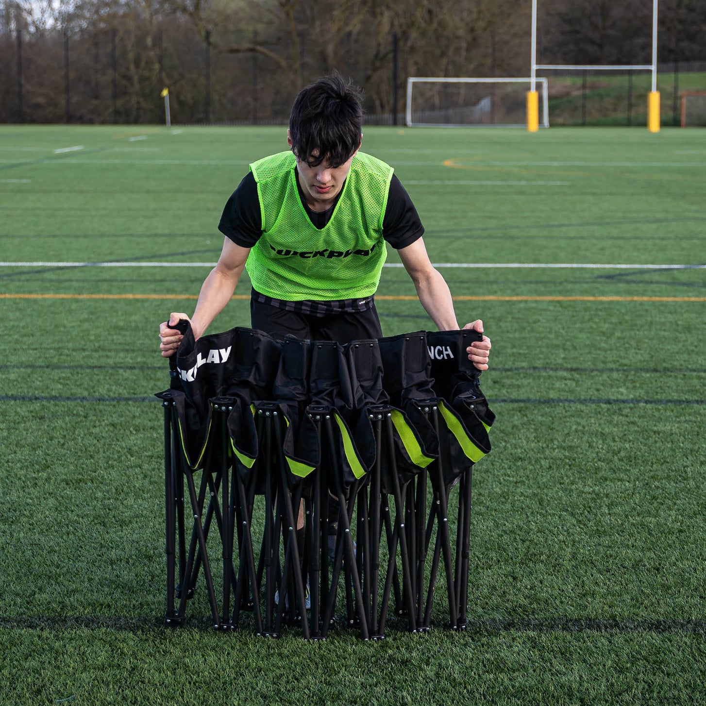 Giocatore con una casacca verde QUICKPLAY che regge la panchina PRO Bench 6 posti parzialmente chiusa su un campo in erba sintetica.