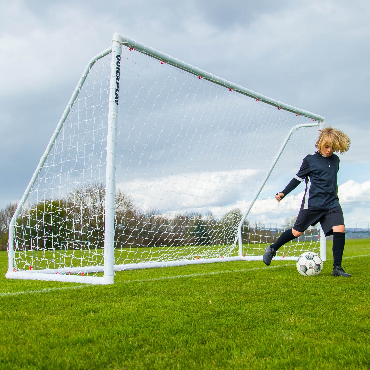 Bambino che dribbla con la palla accanto alla porta Q-FOLD Match 370 x 180 cm su un campo in erba sotto un cielo nuvoloso.