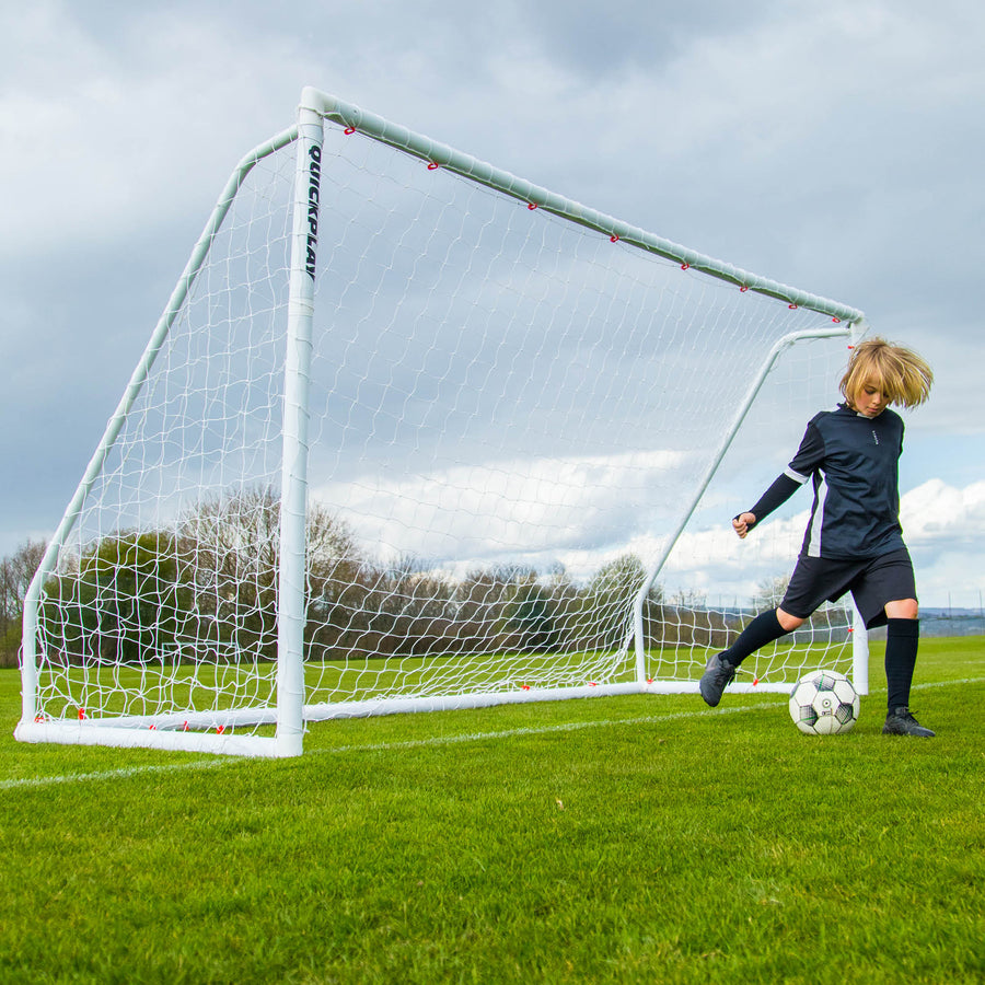 Bambino che dribbla con la palla accanto alla porta Q-FOLD Match 370 x 180 cm su un campo in erba sotto un cielo nuvoloso.