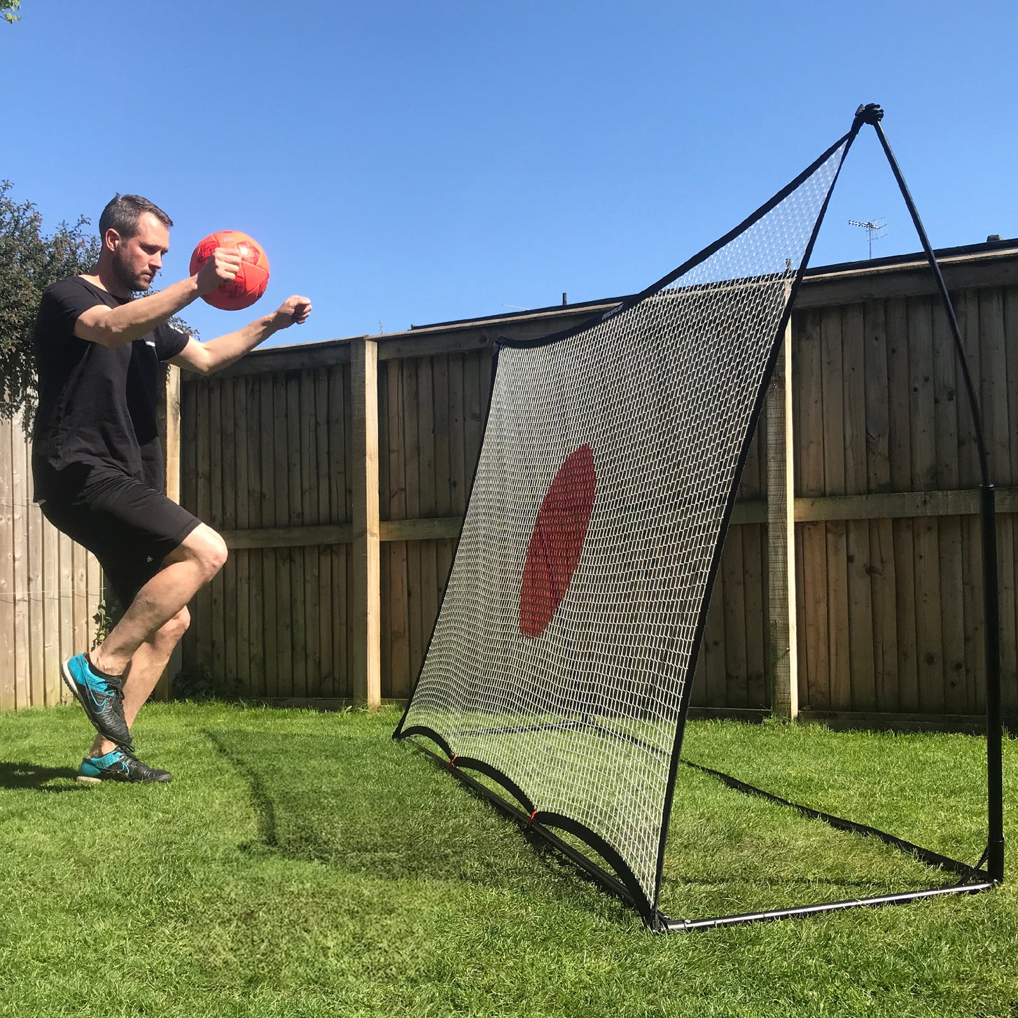 Uomo che tiene una palla rossa accanto al rebounder da calcio SPOT 240 x 150 cm sul prato di un giardino, visto di lato.