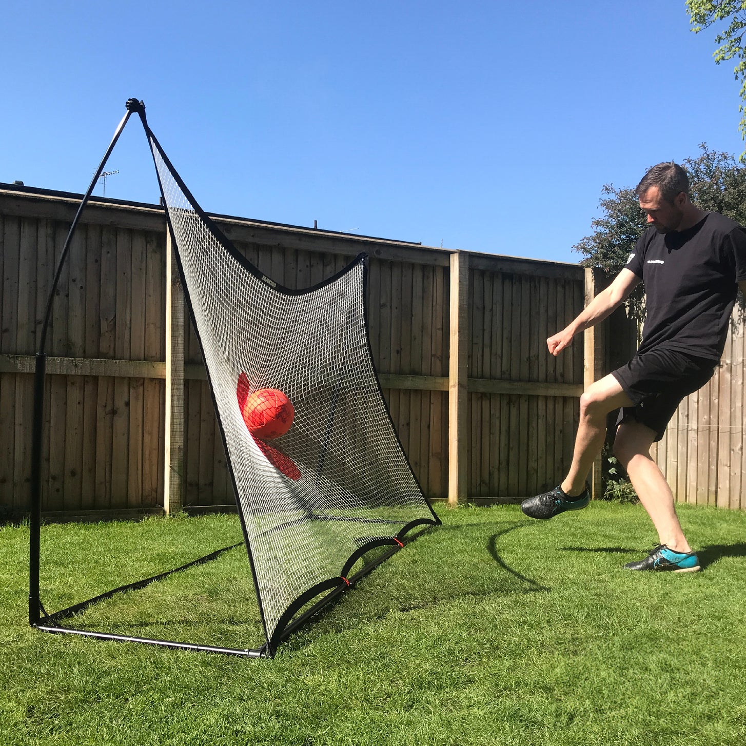 Uomo che calcia una palla rossa nel punto bersaglio rosso del rebounder da calcio SPOT 240 x 150 cm sul prato soleggiato di un giardino.