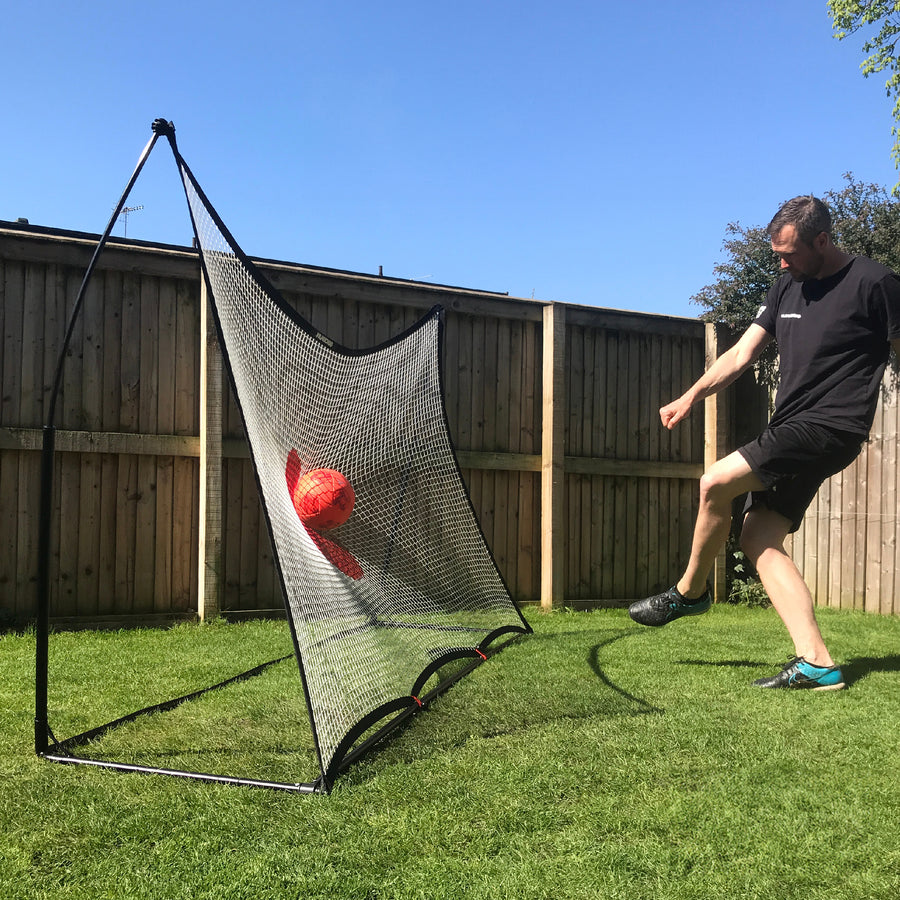 Uomo che calcia una palla rossa nel punto bersaglio rosso del rebounder da calcio SPOT 240 x 150 cm sul prato soleggiato di un giardino.