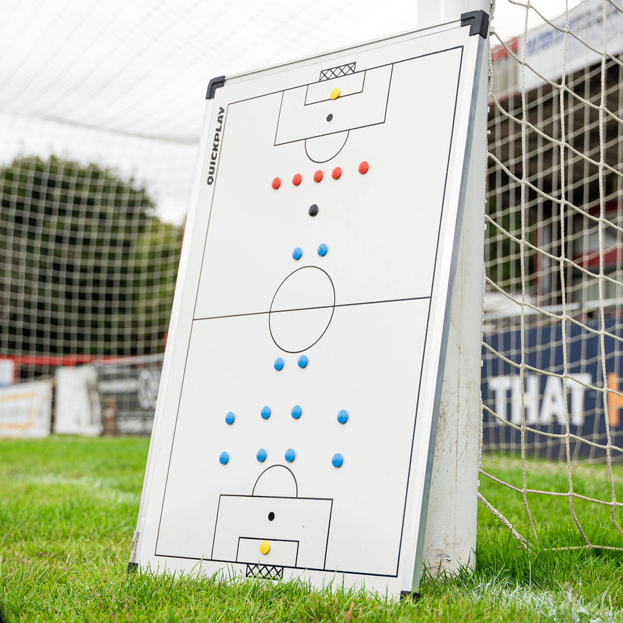 Lavagna tattica magnetica da calcio 60 x 90 cm con lo schema di un campo e magneti colorati, appoggiata a un palo della porta.