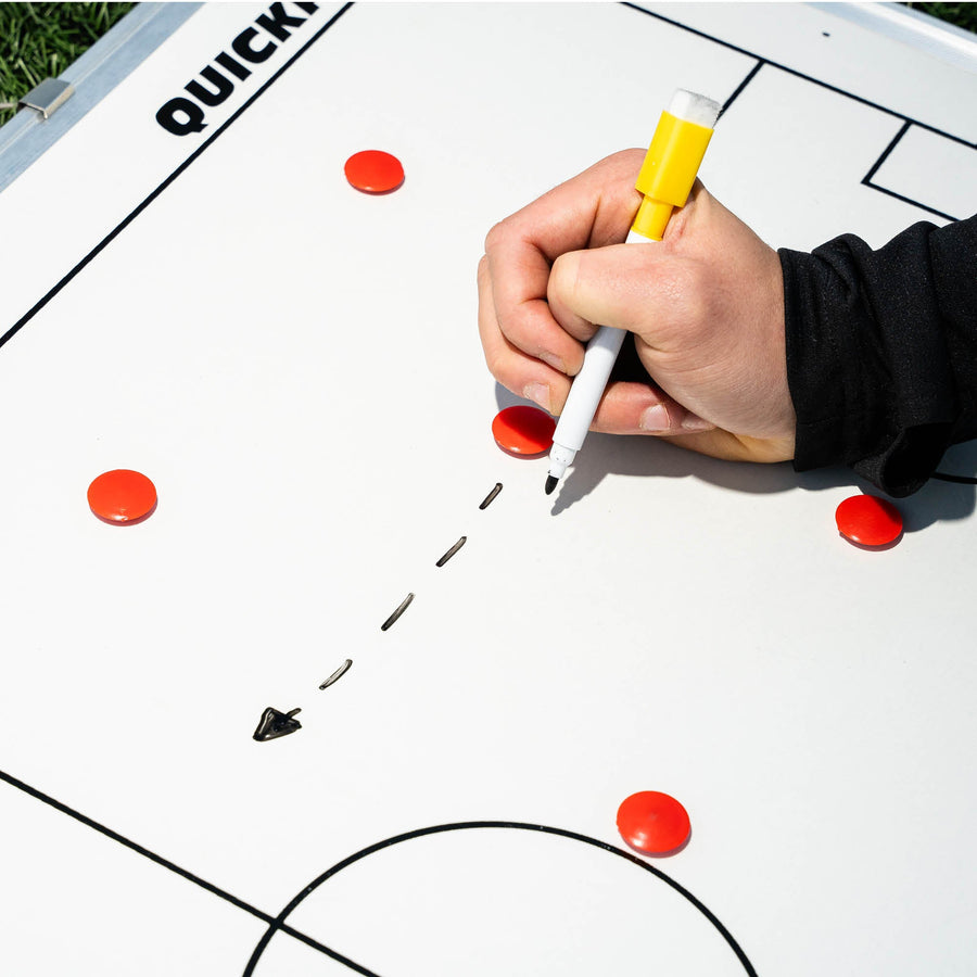 Lavagna tattica magnetica da calcio 60 x 90 cm in uso, con una mano che disegna una freccia tratteggiata con un pennarello giallo tra magneti rossi.