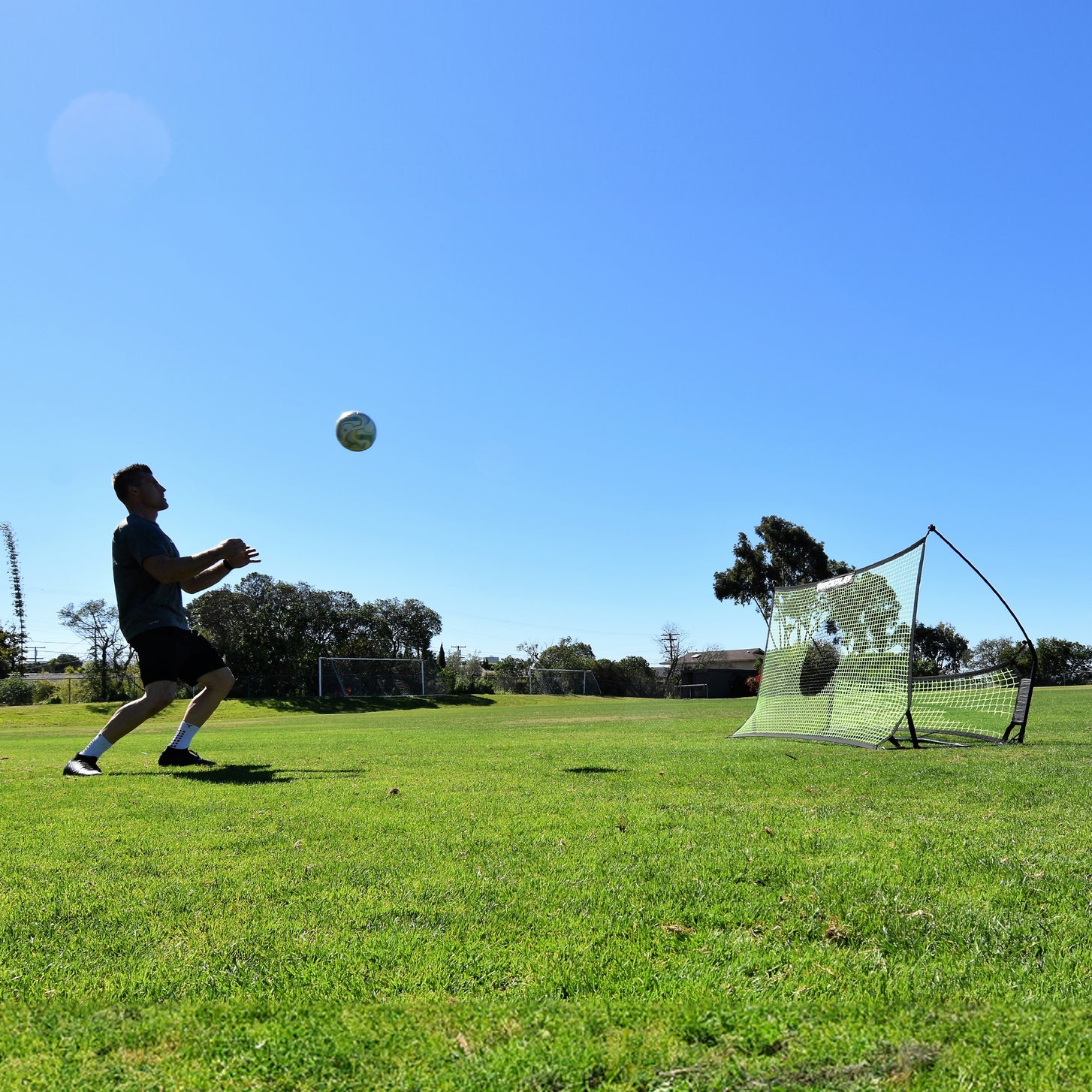 Vista ampia di un giocatore che si prepara a controllare una palla in aria, con il rebounder TEKKERS Trainer dietro di lui sull'erba.